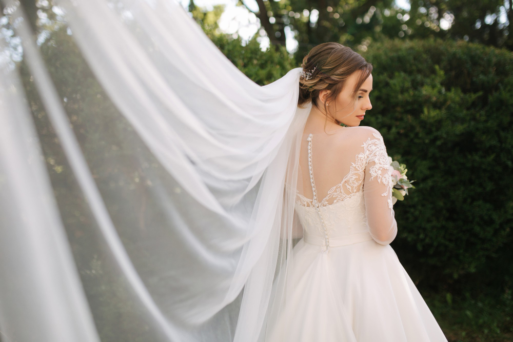 A bride in a long-sleeved lace wedding dress stands outdoors, looking over her shoulder, with her veil flowing behind her.
