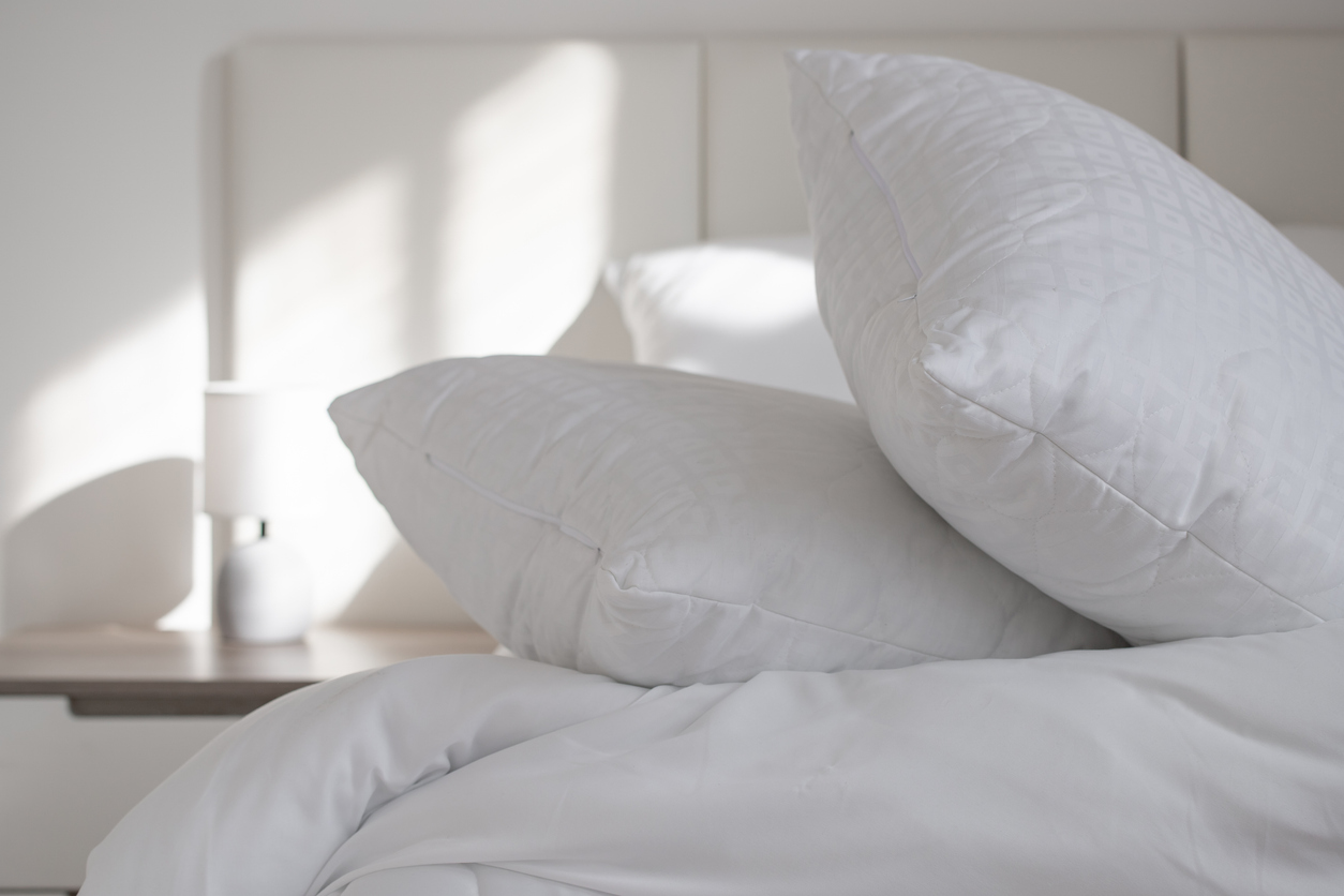 Two white pillows stacked on a neatly made bed in a bright bedroom, with a small lamp and nightstand in the background.