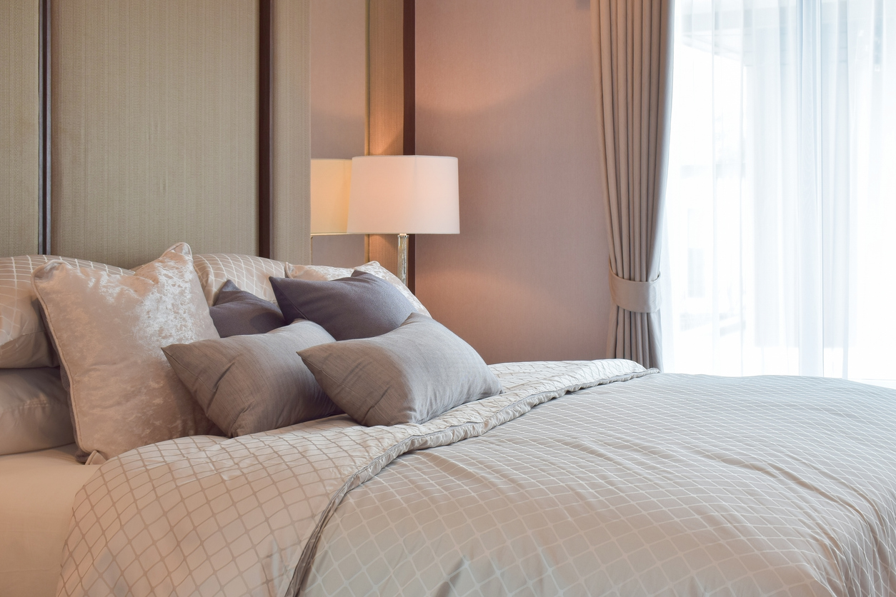 A neatly made bed with multiple gray and white pillows, a patterned comforter, a lamp, and floor-to-ceiling curtains beside a large window letting in natural light.