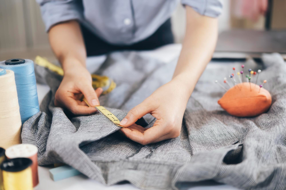 A person measures fabric with a yellow measuring tape on a worktable, surrounded by sewing supplies including spools of thread and a pincushion.