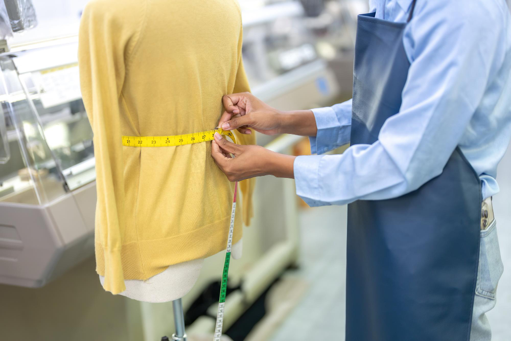 A person wearing an apron uses a measuring tape to take the waist measurement of a mannequin dressed in a yellow sweater.