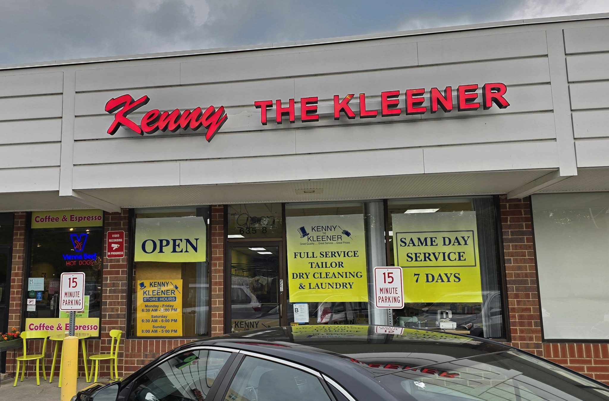 Storefront of “Kenny The Kleener” dry cleaning and laundry shop with bright yellow signage, two 15-minute parking signs, and a black car parked in front.
