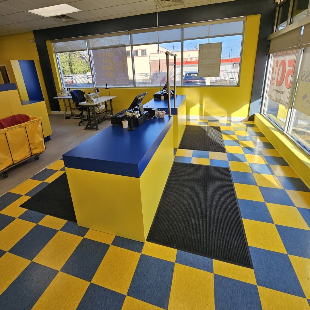 A laundromat interior with blue and yellow checkered floor, blue and yellow counters, black mats, laundry carts, and large windows letting in natural light.