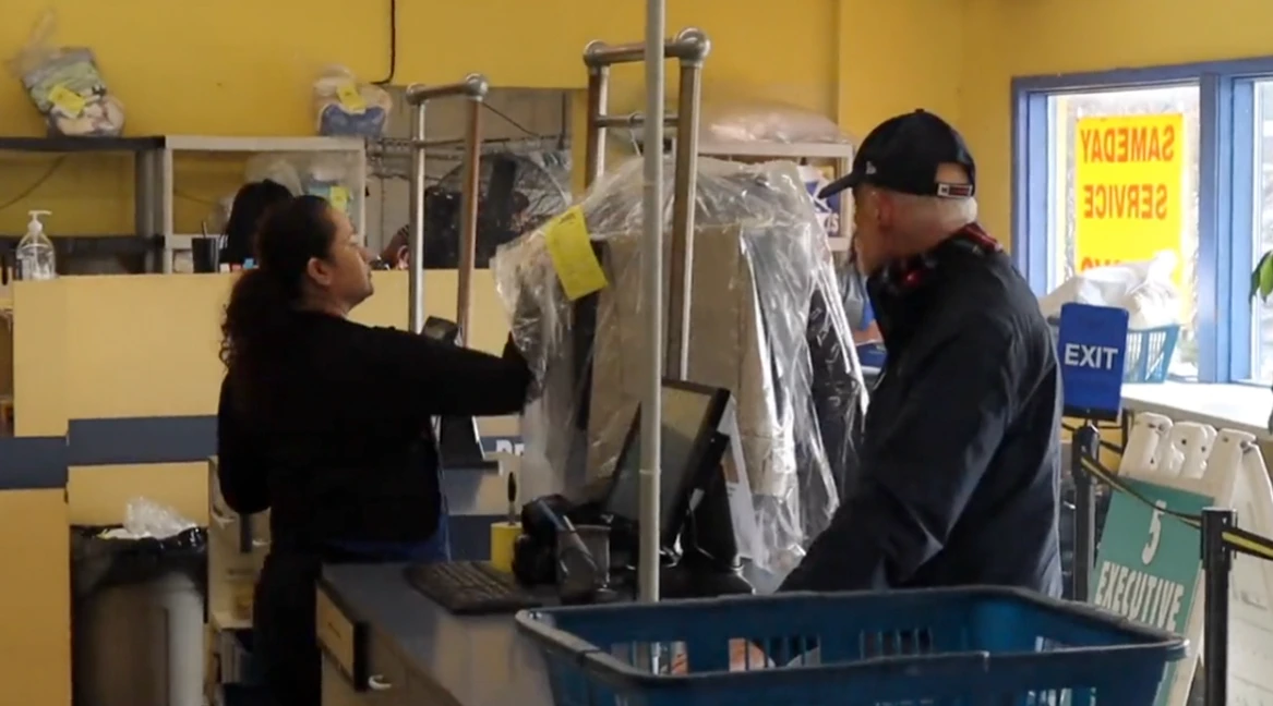 A worker hands over clothes in plastic wrap to a customer at a dry cleaning counter. A blue laundry basket and exit sign are visible in the foreground.