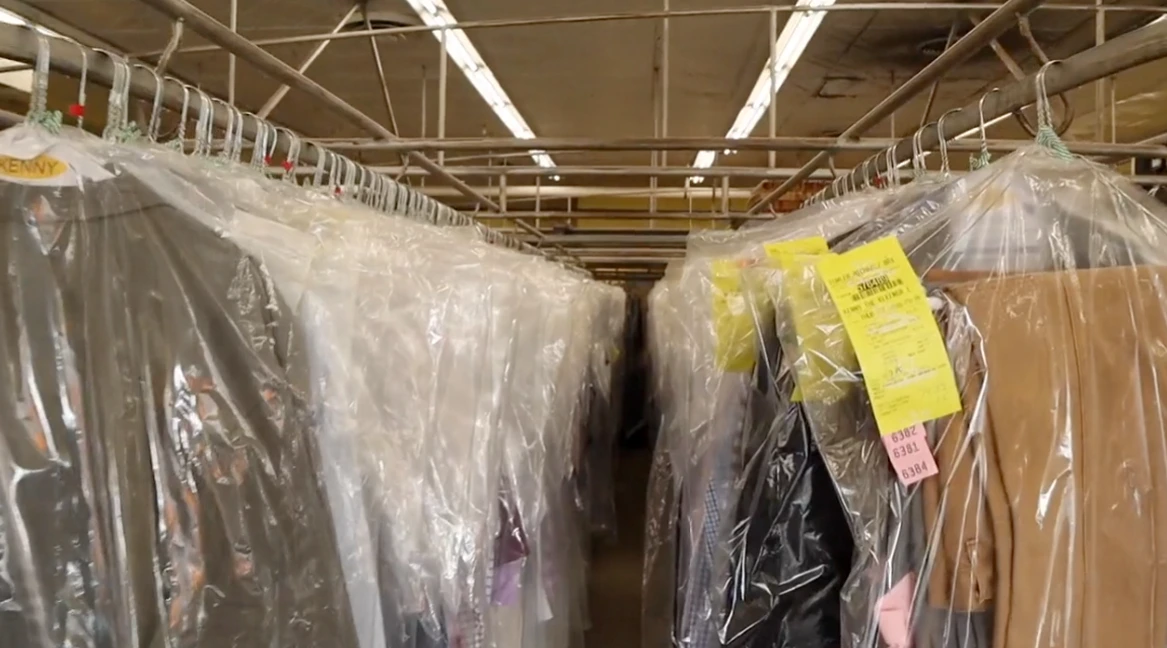Rows of clothes covered in plastic garment bags hang on racks in a dry cleaning facility, with fluorescent lights overhead.