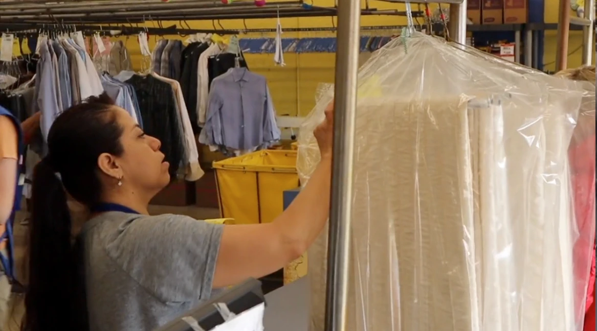 A woman arranges clothes covered in plastic at a dry cleaning facility, with hanging garments and yellow bins visible in the background.