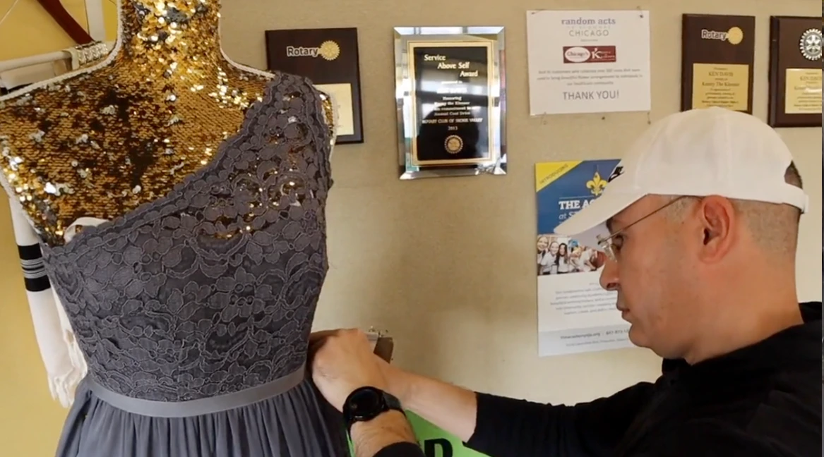A person adjusts the waistline of a gray lace dress on a mannequin, with award plaques and flyers displayed on a bulletin board in the background.