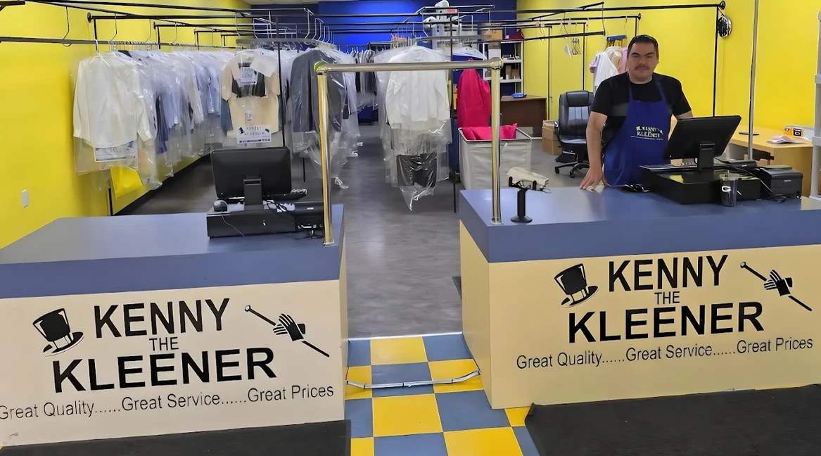 A man stands behind the counter at a dry cleaning shop called "Kenny the Kleener," with clothes hanging in the background and blue and yellow decor.