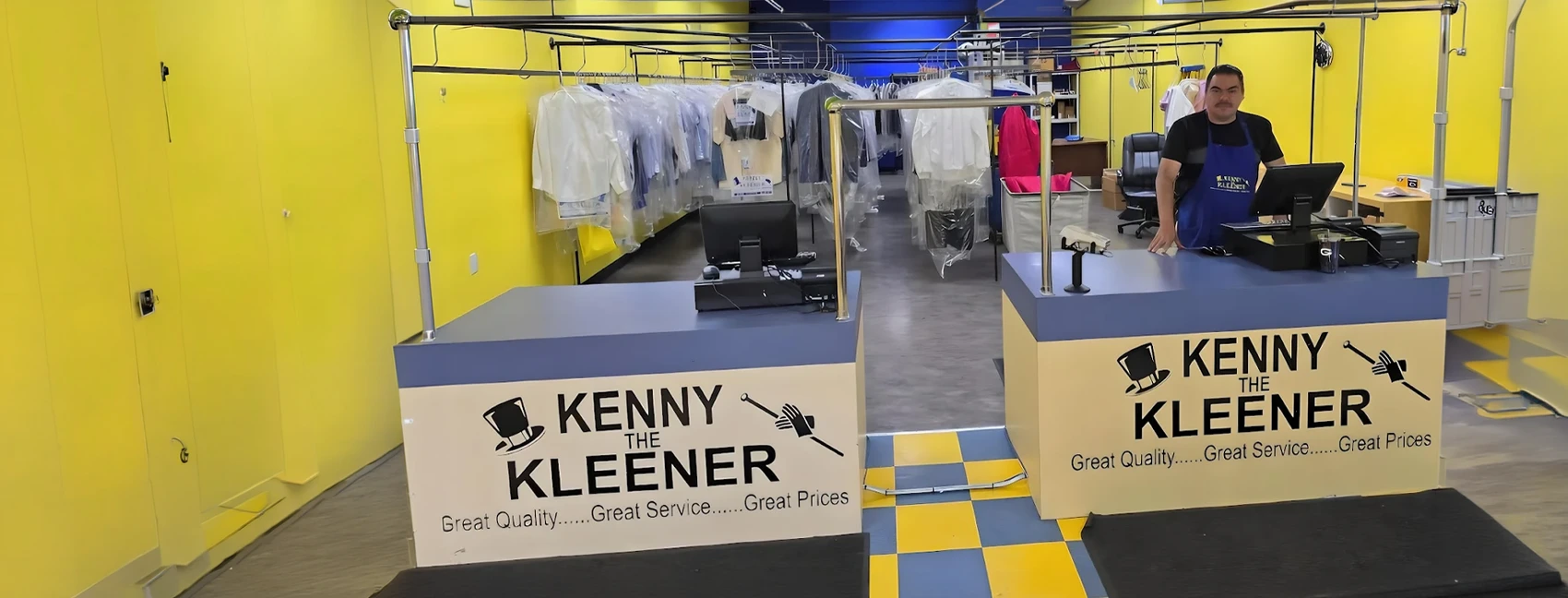 A dry cleaning shop with bright yellow walls, two front counters, clothing racks with garments behind, and an employee standing at one counter.