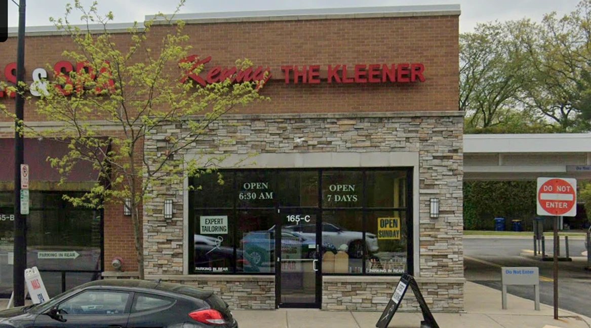 Storefront of "Kerry THE KLEENER" dry cleaners with stone facade, large windows displaying hours, and a car parked in front. A "Do Not Enter" sign is on the corner.