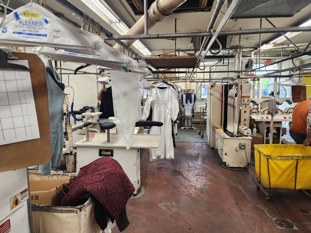 Dry cleaning facility interior with pressing machines, hanging clothes, laundry carts, and workstations visible.