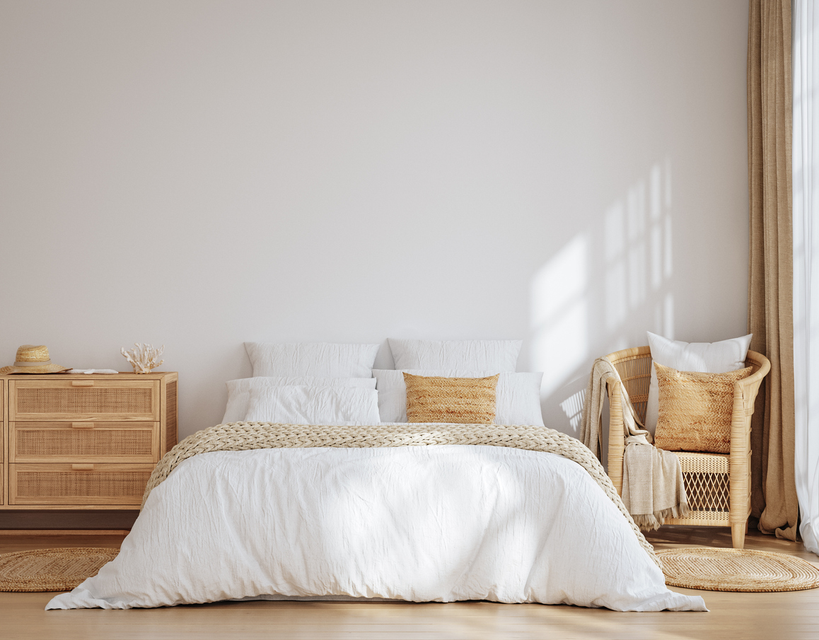 Minimalist bedroom with a white bed, beige and tan pillows, woven chair, wooden dresser, and natural light streaming through a window.