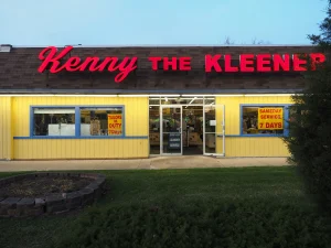 A yellow dry cleaning shop with "Kenny the Kleener" in red letters on the sign and "Sameday Service 7 Days" posted on the windows.
