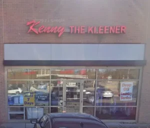 Storefront of "Kenny The Kleener" with large glass windows, signs for dry cleaning, alterations, repairs, and drop-off laundry services visible.