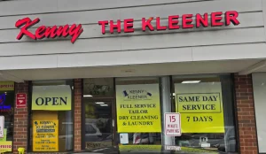 Storefront of "Kenny the Kleener" dry cleaning shop with signs for full service, tailoring, same day service, and an "OPEN" sign in the window.