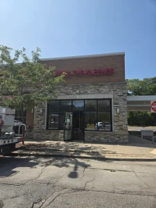 A brick and stone commercial building with a large window and a partially obscured red sign reading "THE KLEENER", facing a street with a tree and a parked truck.