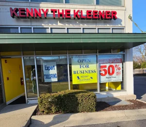Storefront of "Kenny the Kleener" with signs for expert tailoring, open for business, and a grand opening 50% off promotion displayed in the windows.