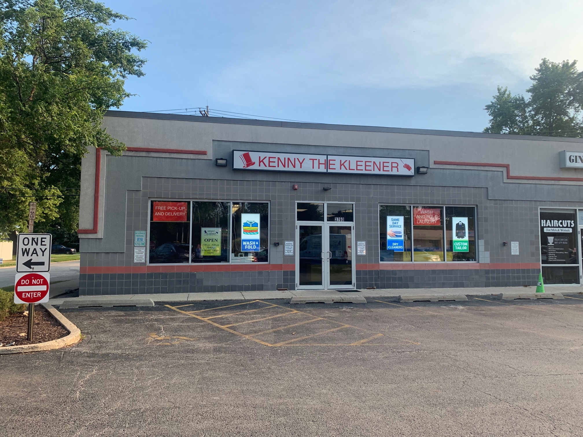 A dry cleaning store named "Kenny the Kleener" with large front windows, parking spaces, and multiple service signs by the entrance.